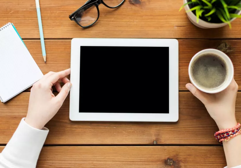 Aerial view of a desk with person's hands holding tablet computer and cup of coffee