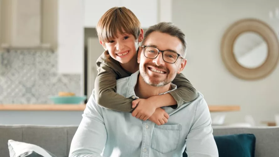 Boy with arms around a man, smiling to camera, inside a living room