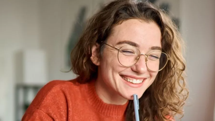 Woman smiling, with pen to chin, appears to be looking at a computer screen