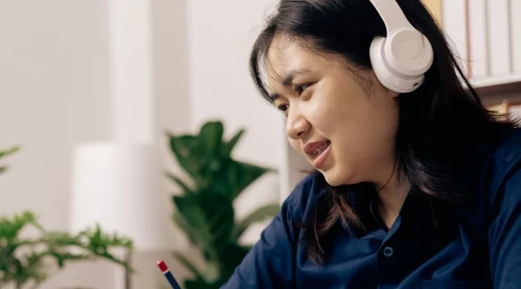 Woman wearing headphones and writing in notebook while looking at a laptop, in a home office environment
