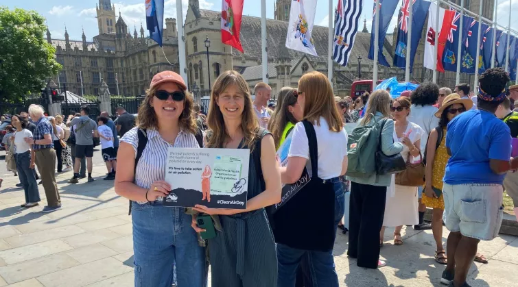 Two women outside Palace of Westminster and crowd holding a sign that reads "We treat patients suffering health harms of polluted air every day. It's time to take action on air pollution. #CleanAirDay"