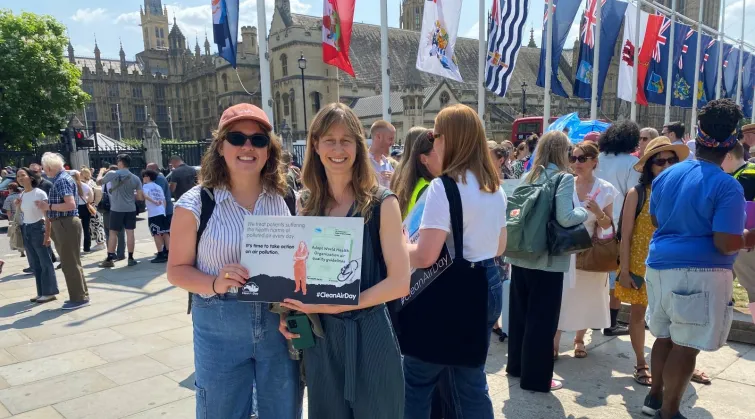 Two women outside Palace of Westminster and crowd holding a sign that reads "We treat patients suffering health harms of polluted air every day. It's time to take action on air pollution. #CleanAirDay"