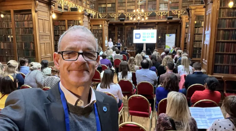 Photo: Steve Turner at a conference, in a library lined with books, with a presenter at front and seated delegates