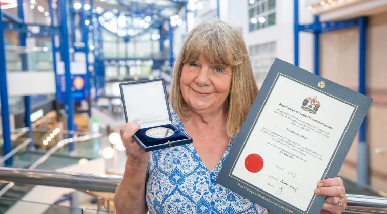 Julia Donaldson at a conference centre. In one hand she holds the College Medal, a silver disc in a case, and in the other hand a certificate 
