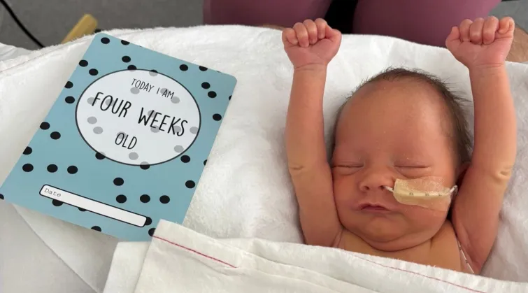 Small baby in hospital bed with arms in air, next to a card reading 'Today I am four weeks old'