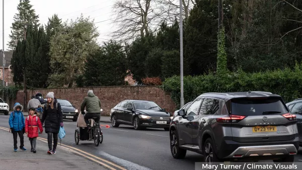 Image depicts woman and two children walking along a street next to a cyclist and cars