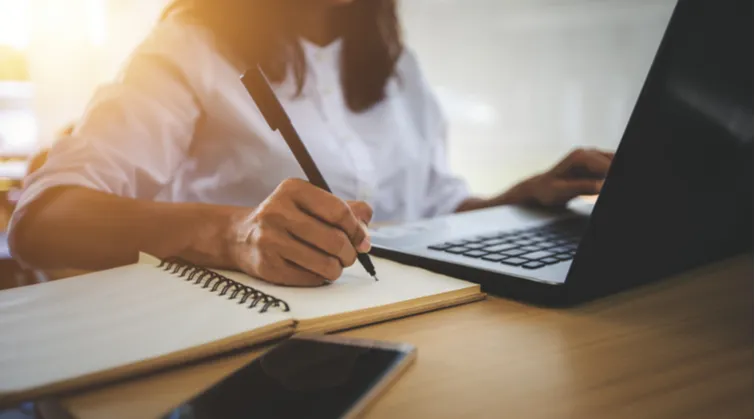 Closeup of person at laptop with notepad and pen, sunshine behind