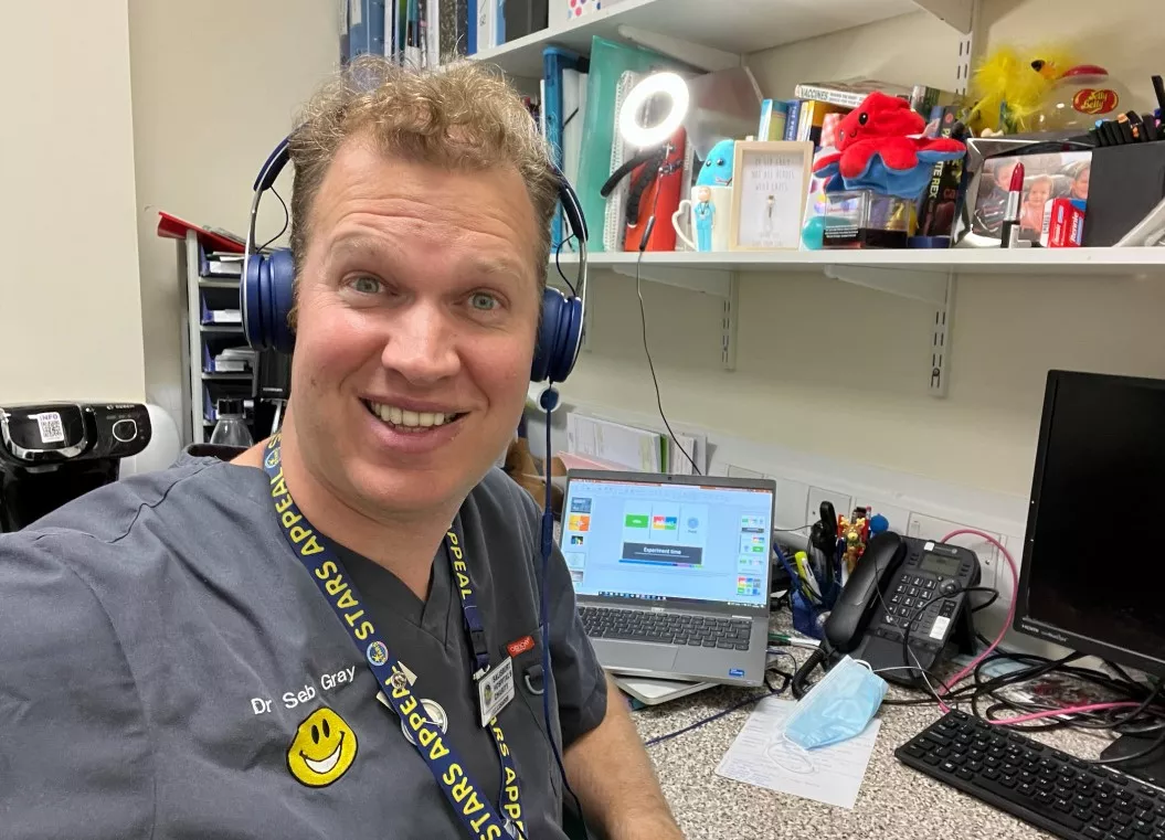 Seb Gray at his work computer, wearing headphones, a smiley face embroidered on his scrubs