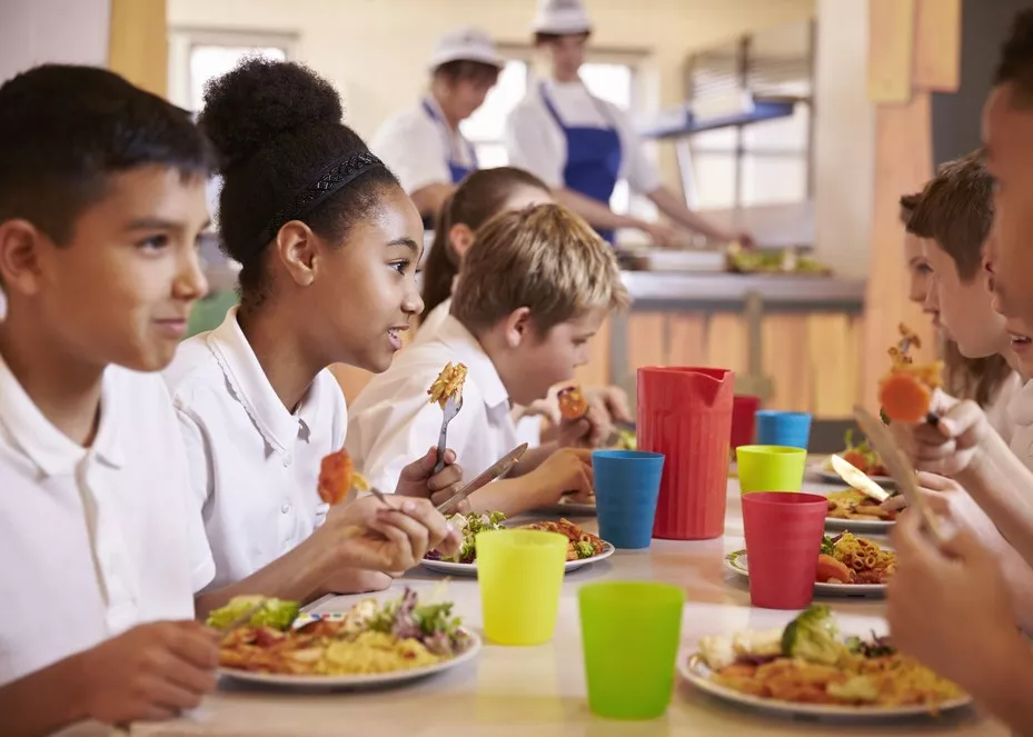 Group of primary school students eating lunch at a table, with cafeteria staff behind