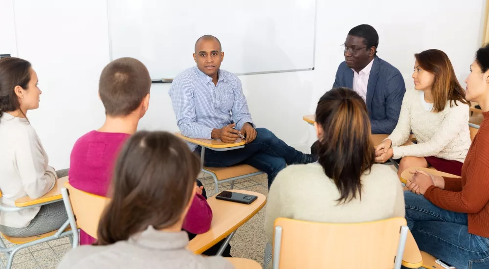 Group of people sitting at individual desks, looking at instructor, with whiteboard behind