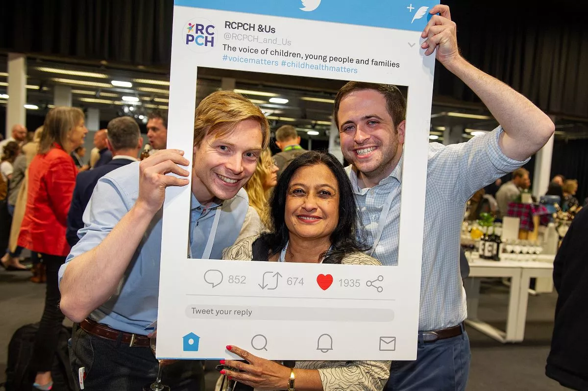 Three people look through a large cardboard frame, reading RCPCH &Us: the voice of children, young people and families, shaped like a Twitter post  