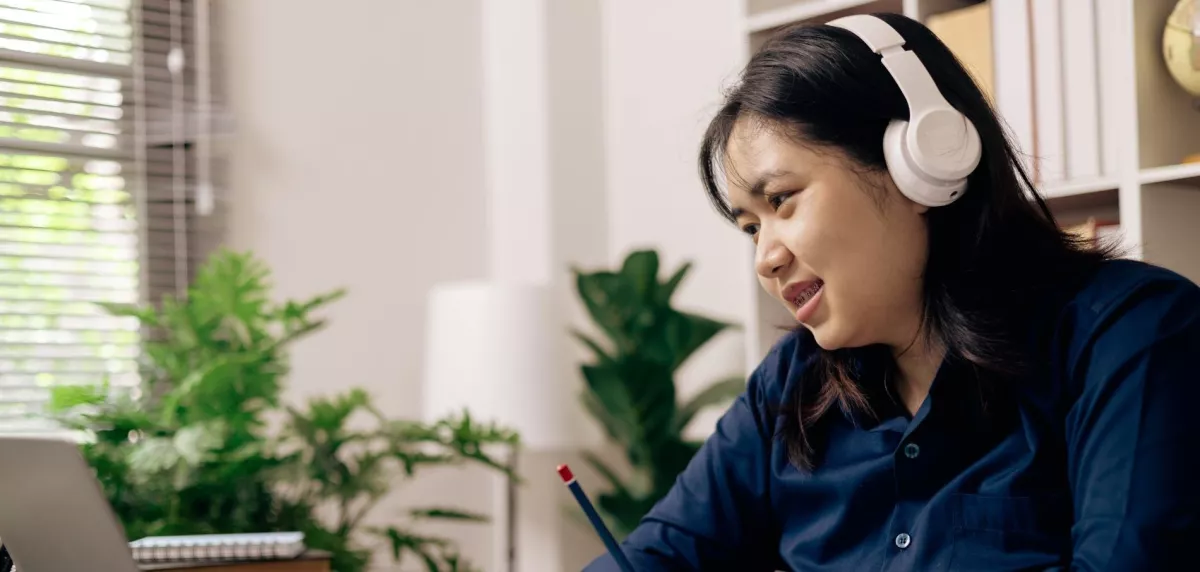 Woman wearing headphones and writing in notebook while looking at a laptop, in a home office environment