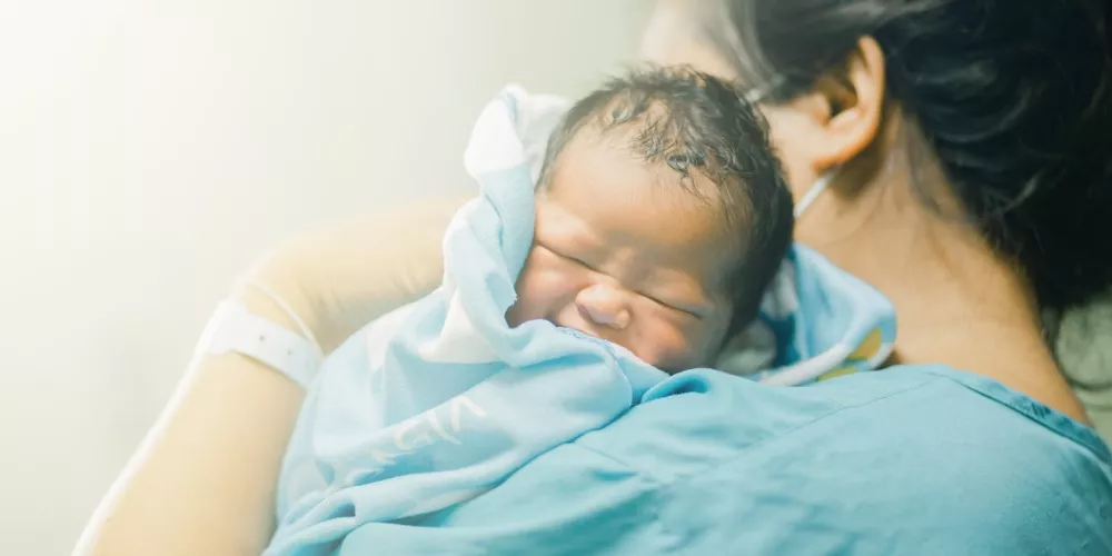 Small baby, being held by adult wearing scrubs and mask