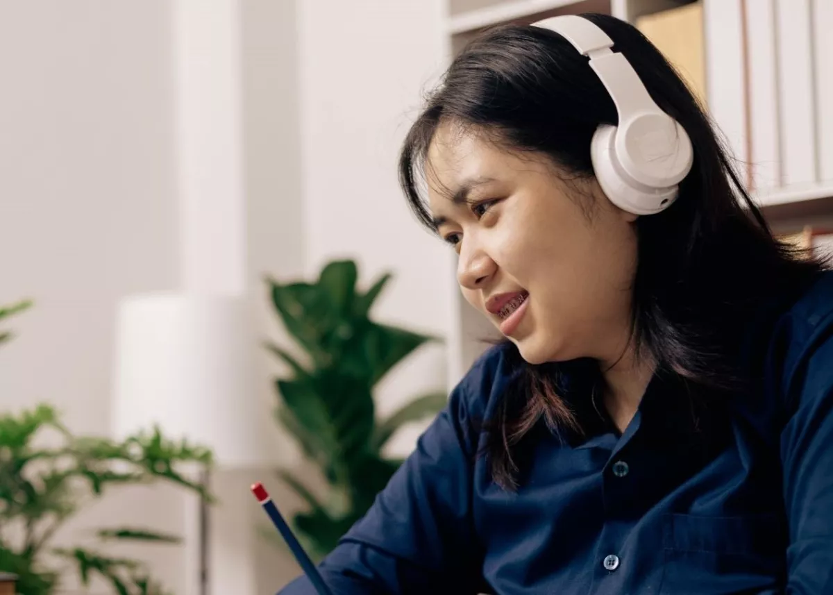 Woman wearing headphones and writing in notebook while looking at a laptop, in a home office environment