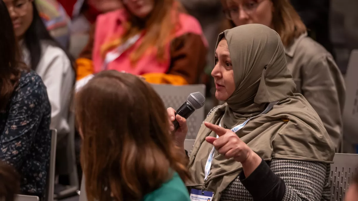 Woman speaking into a microphone, seated, with other people around her looking on