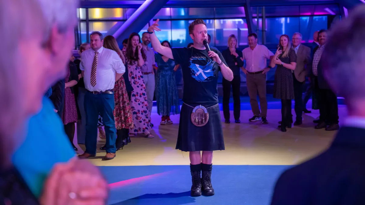 Main in kilt and sporran, holding microphone, surrounded by others