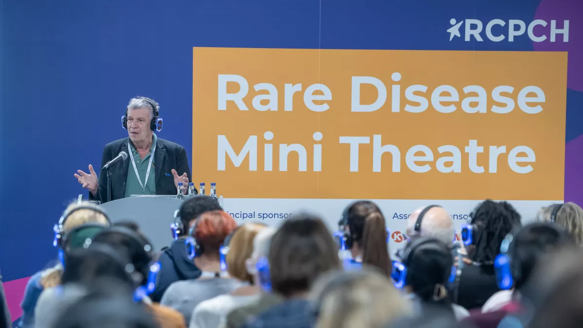 Speaker with audience wearing headphones, sign in background reads RCPCH Rare Disease Mini Theatre 