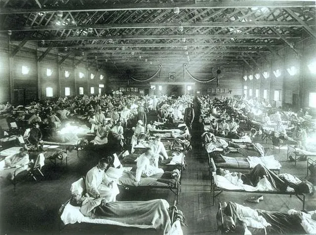An image showing an early 20th century hospital ward set up in a warehouse with long rows of beds, and nurses tending the sick