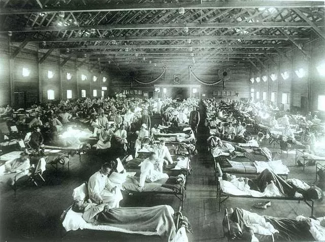 An image showing an early 20th century hospital ward set up in a warehouse with long rows of beds, and nurses tending the sick