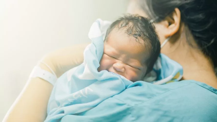 Small baby, being held by adult wearing scrubs and mask