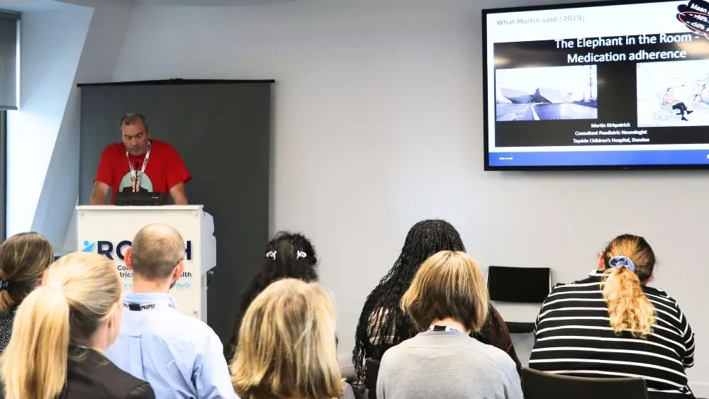 Man presenting from podium to an audience - with slide reading 'Elephant in the room, medicines adherence'