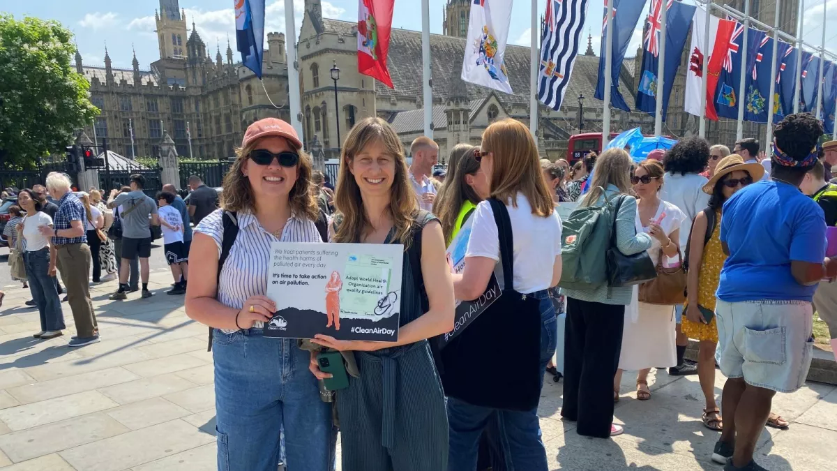 Two women outside Palace of Westminster and crowd holding a sign that reads "We treat patients suffering health harms of polluted air every day. It's time to take action on air pollution. #CleanAirDay"