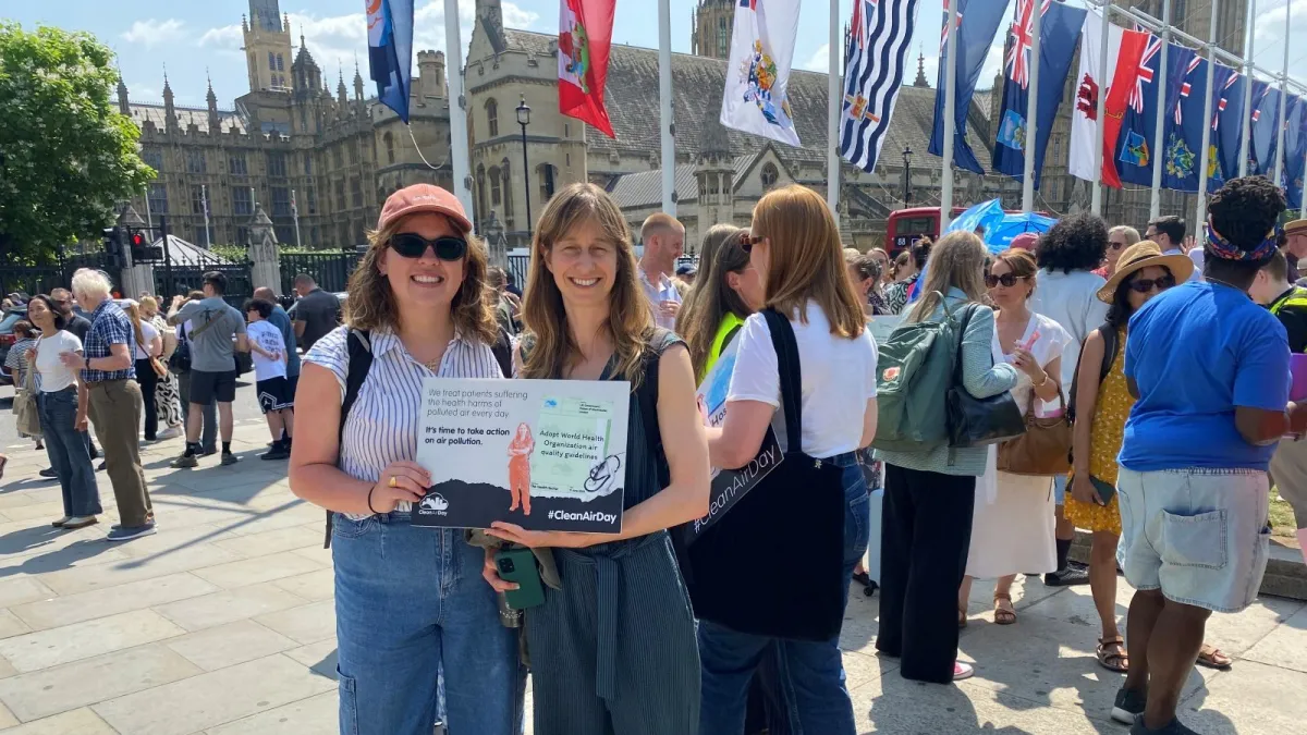 Two women outside Palace of Westminster and crowd holding a sign that reads "We treat patients suffering health harms of polluted air every day. It's time to take action on air pollution. #CleanAirDay"