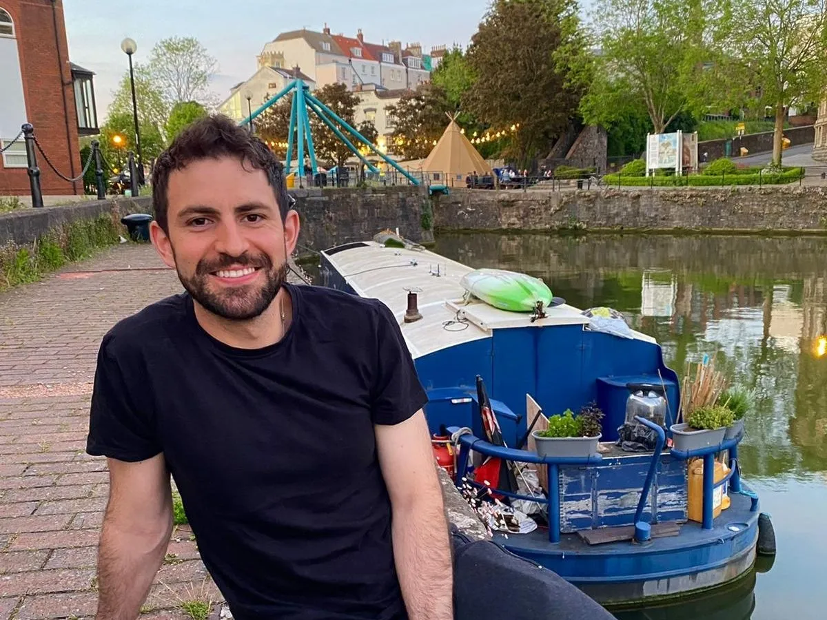 Peter Eriksen, sitting by canal with a houseboat and bridge behind him