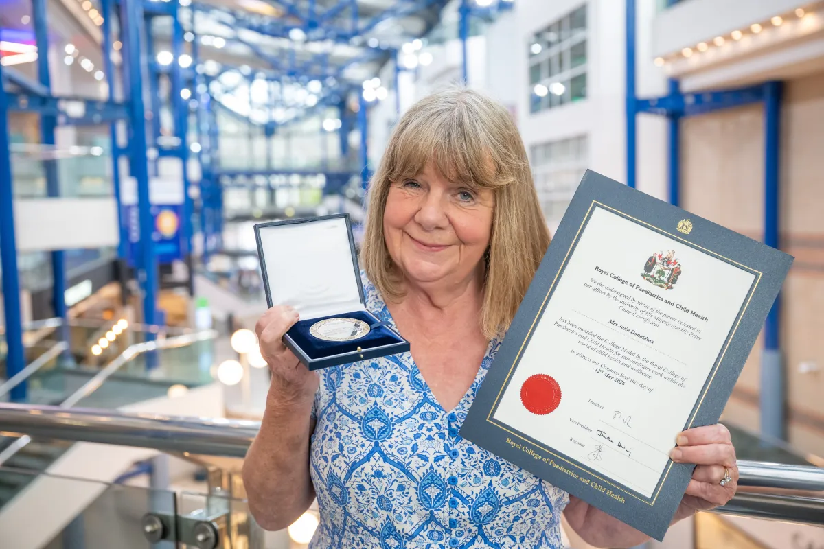 Julia Donaldson at a conference centre. In one hand she holds the College Medal, a silver disc in a case, and in the other hand a certificate 
