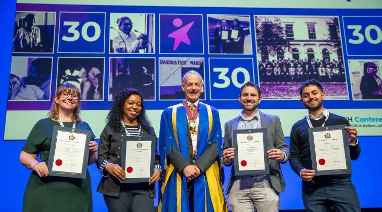 The four winners of the PAFTA display their certificates while standing next to RCPCH President on a stage, with a backdrop showing RCPCH 30 years and photos