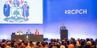 A stage with a man at podium and others at a table; with RCPCH and a crest displaying on screen; a large audience watches
