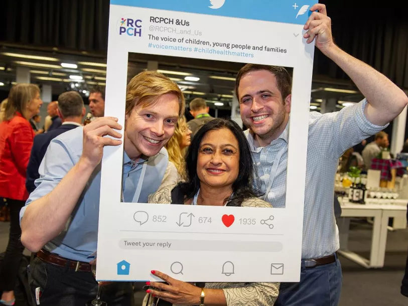 Three people look through a large cardboard frame, reading RCPCH &Us: the voice of children, young people and families, shaped like a Twitter post  