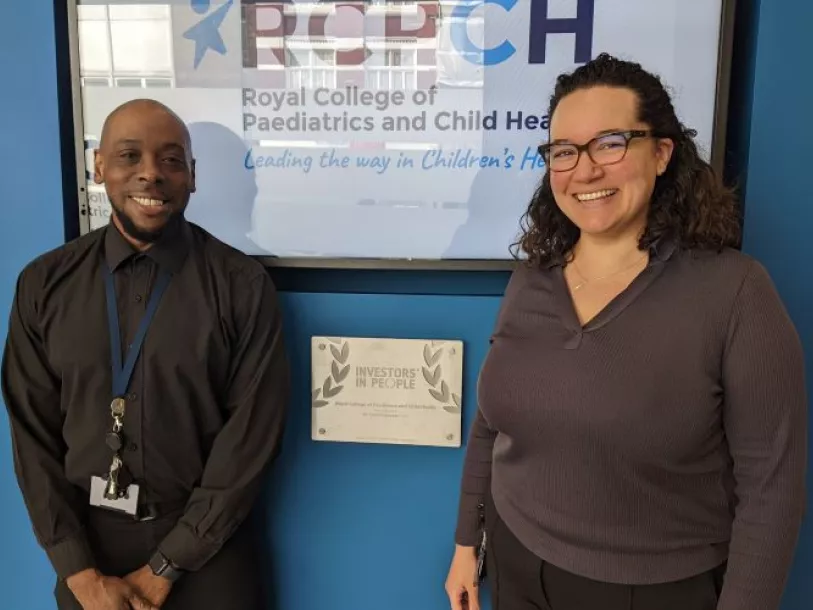Two staff members stand in front of RCPCH sign and Investors in People silver award plaque 