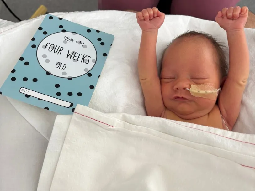 From the cover of NNAP summary report on 2025 data; Photo courtesy of Morag McInnes Small baby in hospital bed with arms in air, next to a card reading 'Today I am four weeks old'